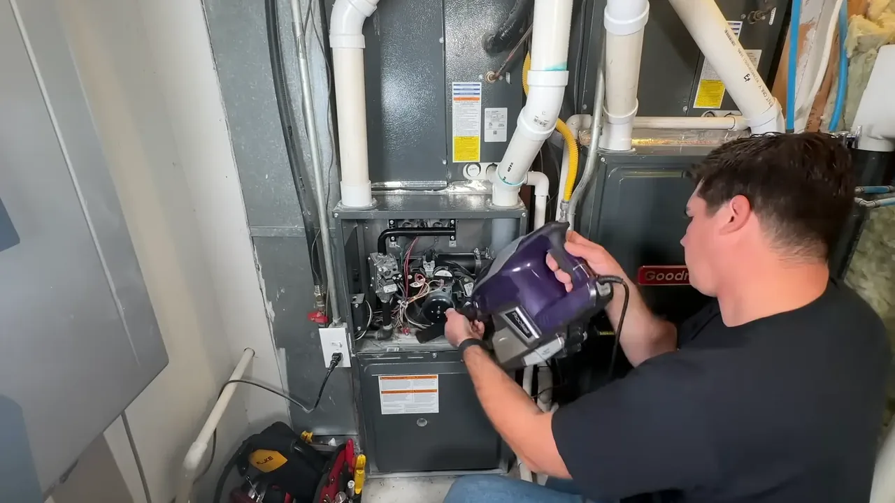 Technician using a handheld vacuum to clean the inside of an open furnace, with a tool bag on the floor beside it